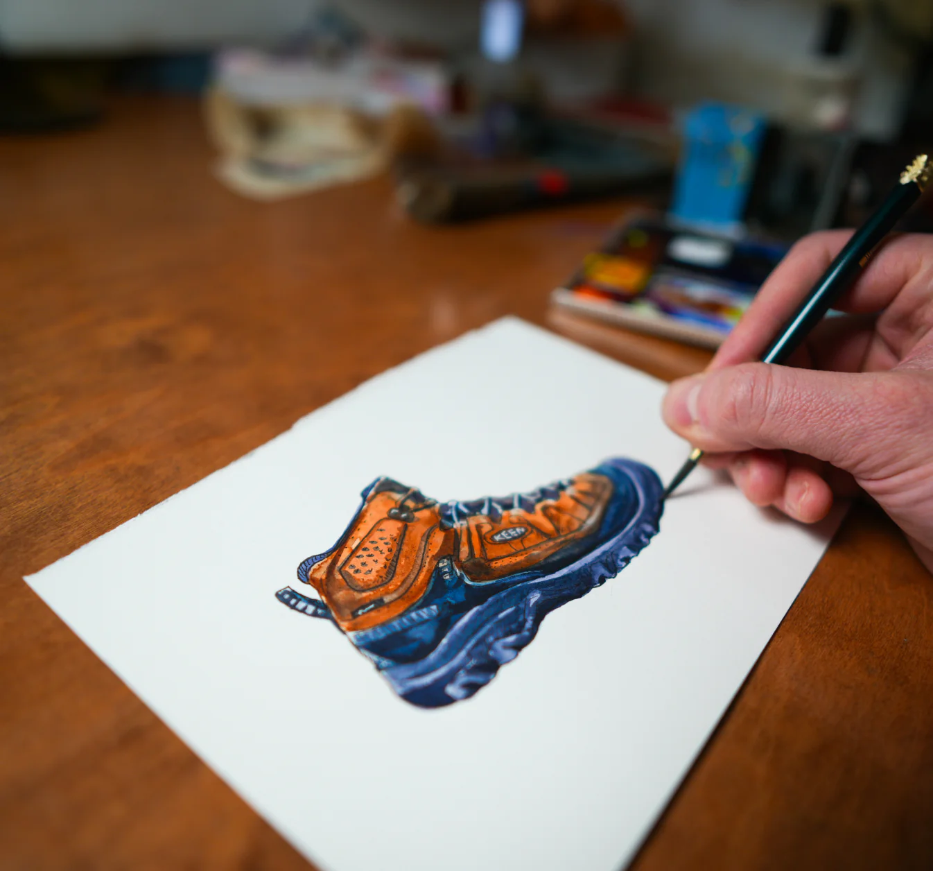 A man's hand holding a paintbrush and painting a Keen Targhee hiking boot on white paper