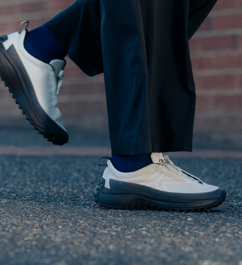 A person walking along the street, wearing white KEEN sneakers with black soles.