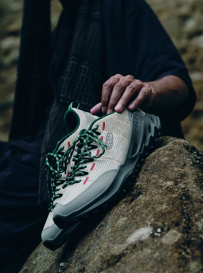 A person on a rock holds sneakers featuring green laces and accent stitching, dressed in casual clothing in outdoor setting.